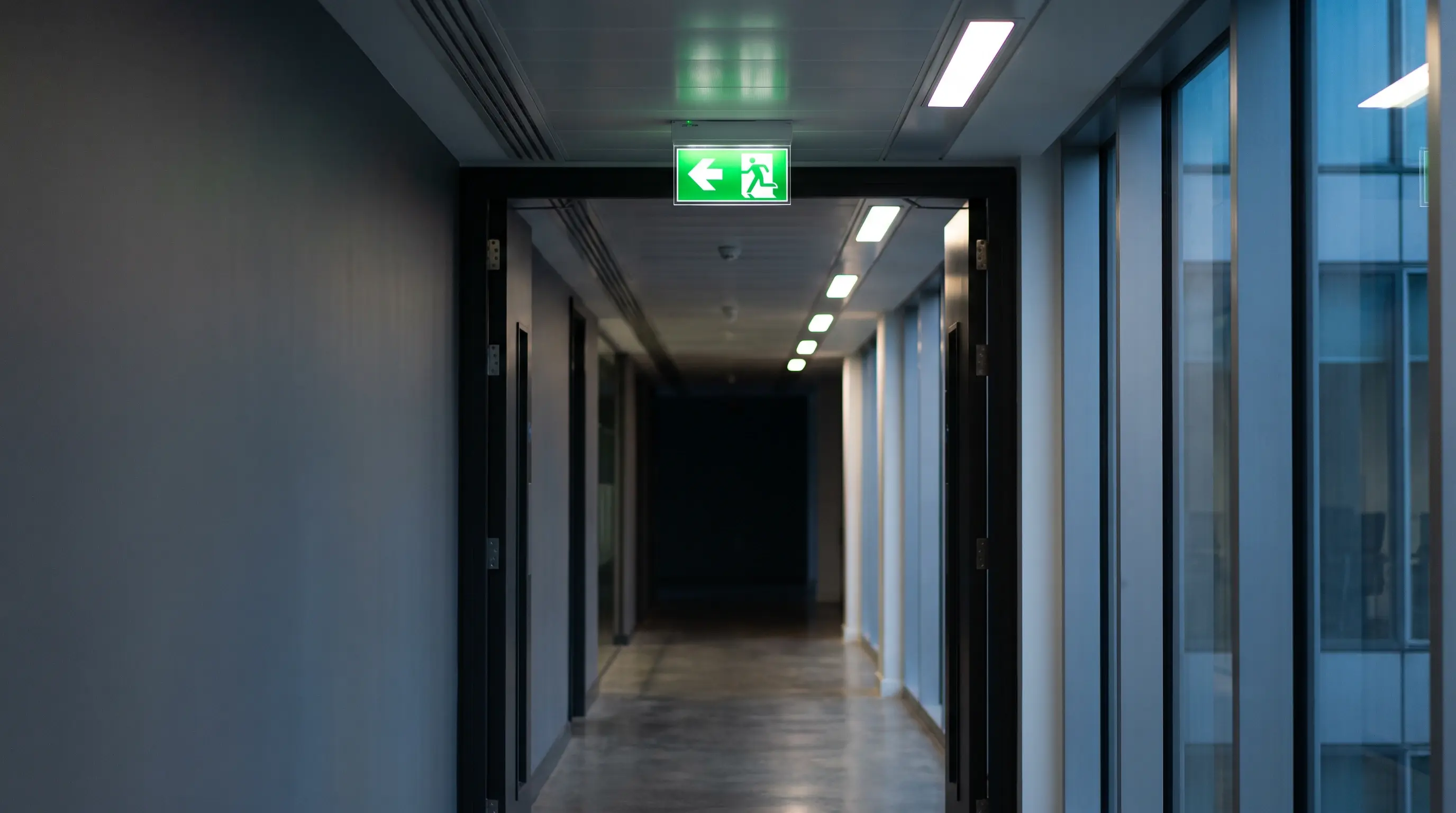 Office corridor illuminated by ceiling-mounted emergency lighting, with a green exit sign hanging above the doorway pointing toward the fire escape route